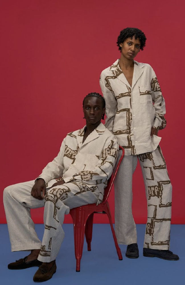 Two models wearing cream linen shirts with abstract print pattern and coordinating pants against vibrant pink and blue background